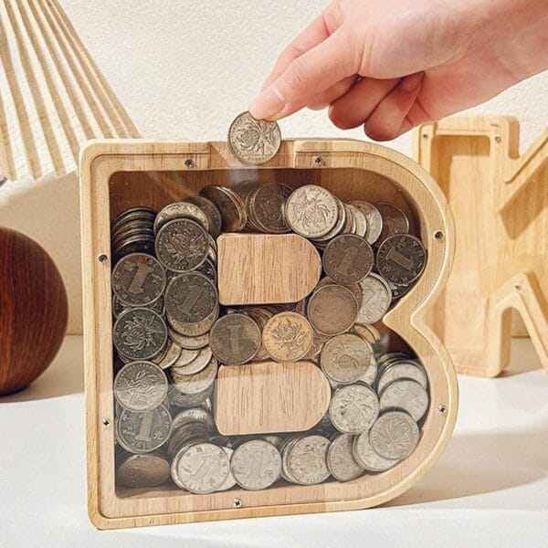Wooden letter B piggy bank filled with coins, hand adding more. Savings concept.