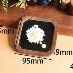 Square wooden jewelry display case with a white floral brooch and pearl pendant on a black velvet base.