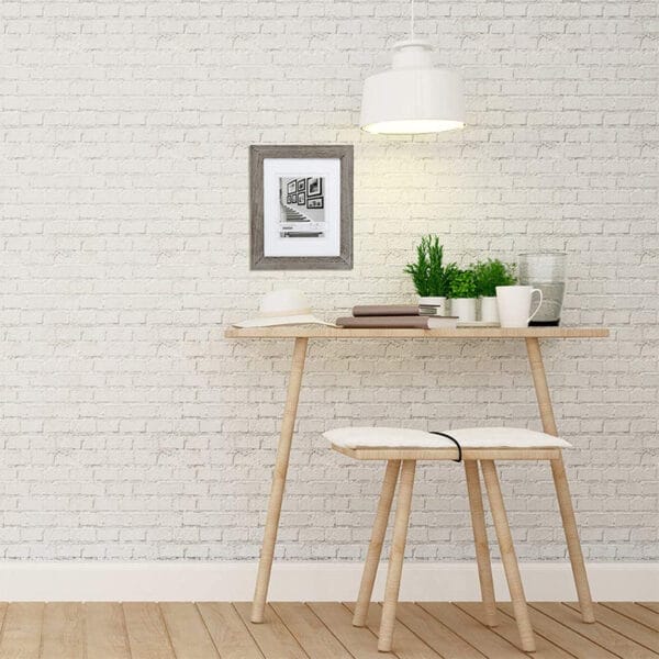 Gray picture frame on white brick wall above a wooden desk and stool.