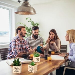 Four people meet at a table with succulent plant gifts in wooden pots, each inscribed with "Thank you for being awesome.