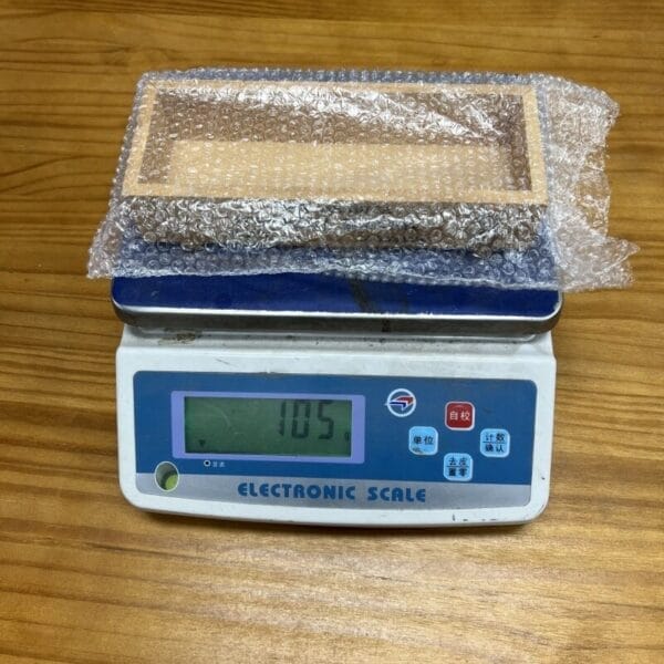 Wooden tray wrapped in bubble wrap on a scale, weighing 105 units. Packaging for shipping.